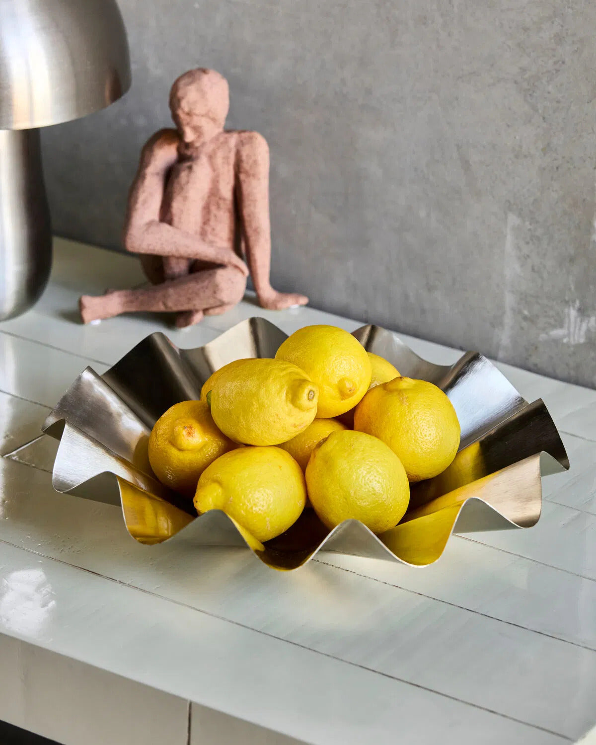 Brushed silver flood display bowl sits on a counter filled with lemons. In the background is a clay figure and a silver lamp.