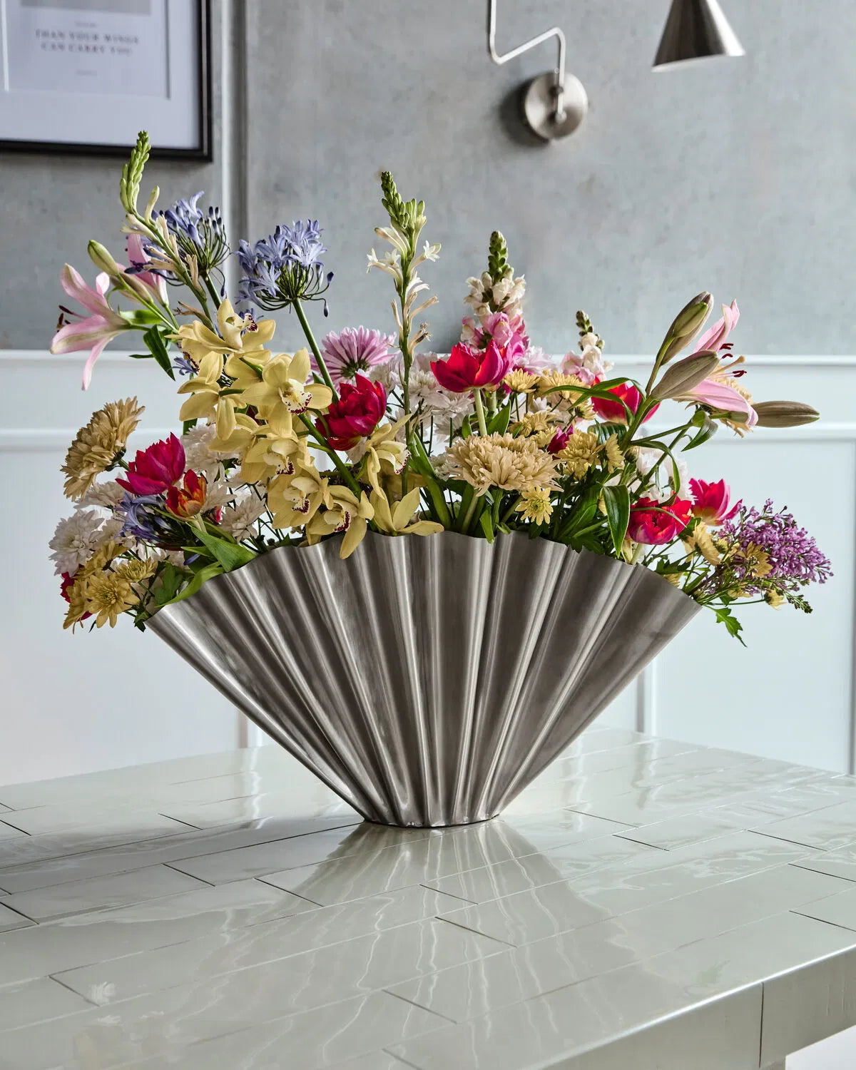 A large brushed silver Flood vase filled with flowers, standing on a tiled table.