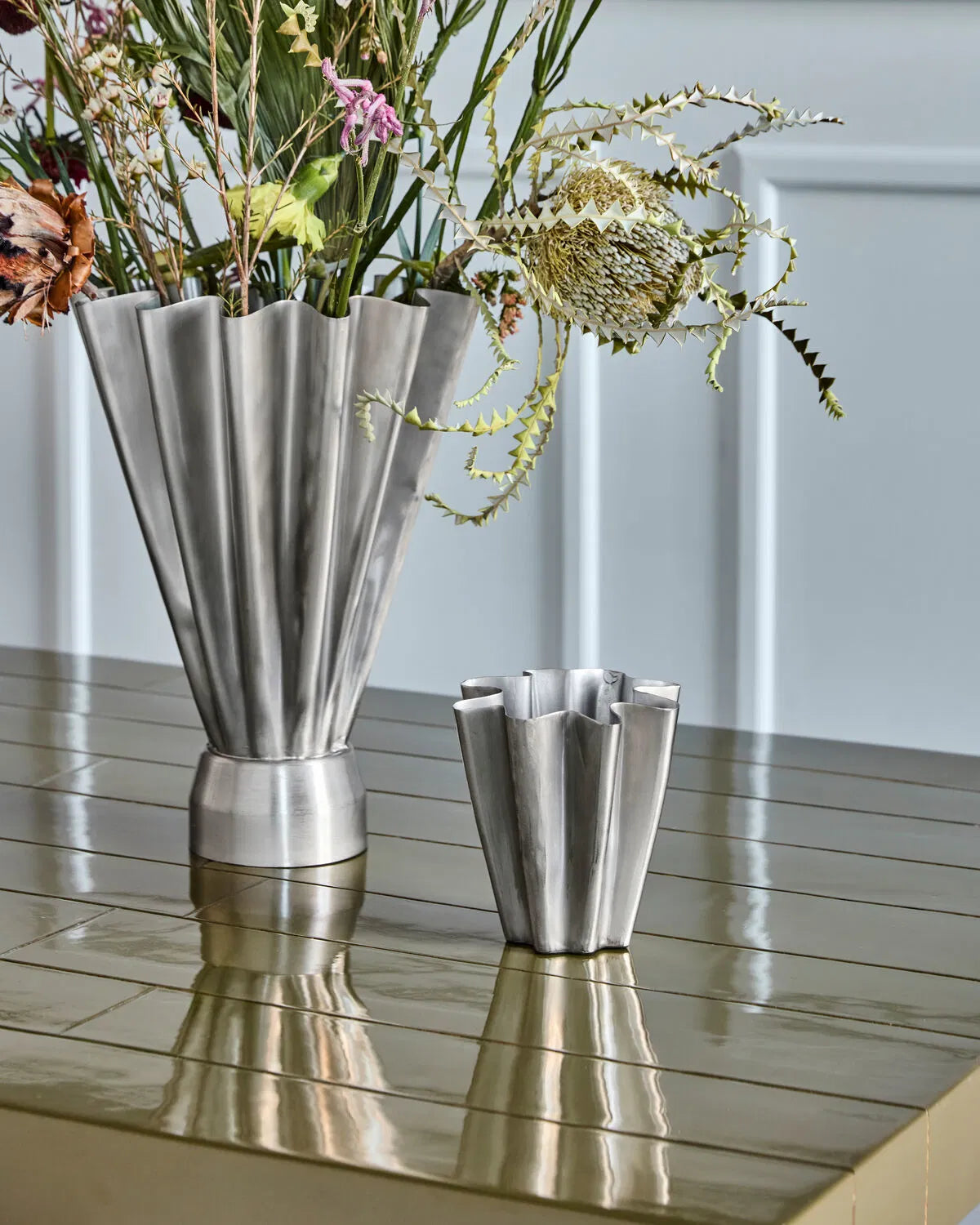 A brushed silver, small flood vase stands on a table next to a larger size of the same design. The larger vase is filled with flowers, but the smaller vase is not.
