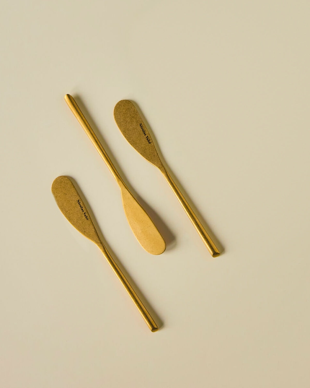 Three Nicolas Vahé butter knives in antique gold on a cream background