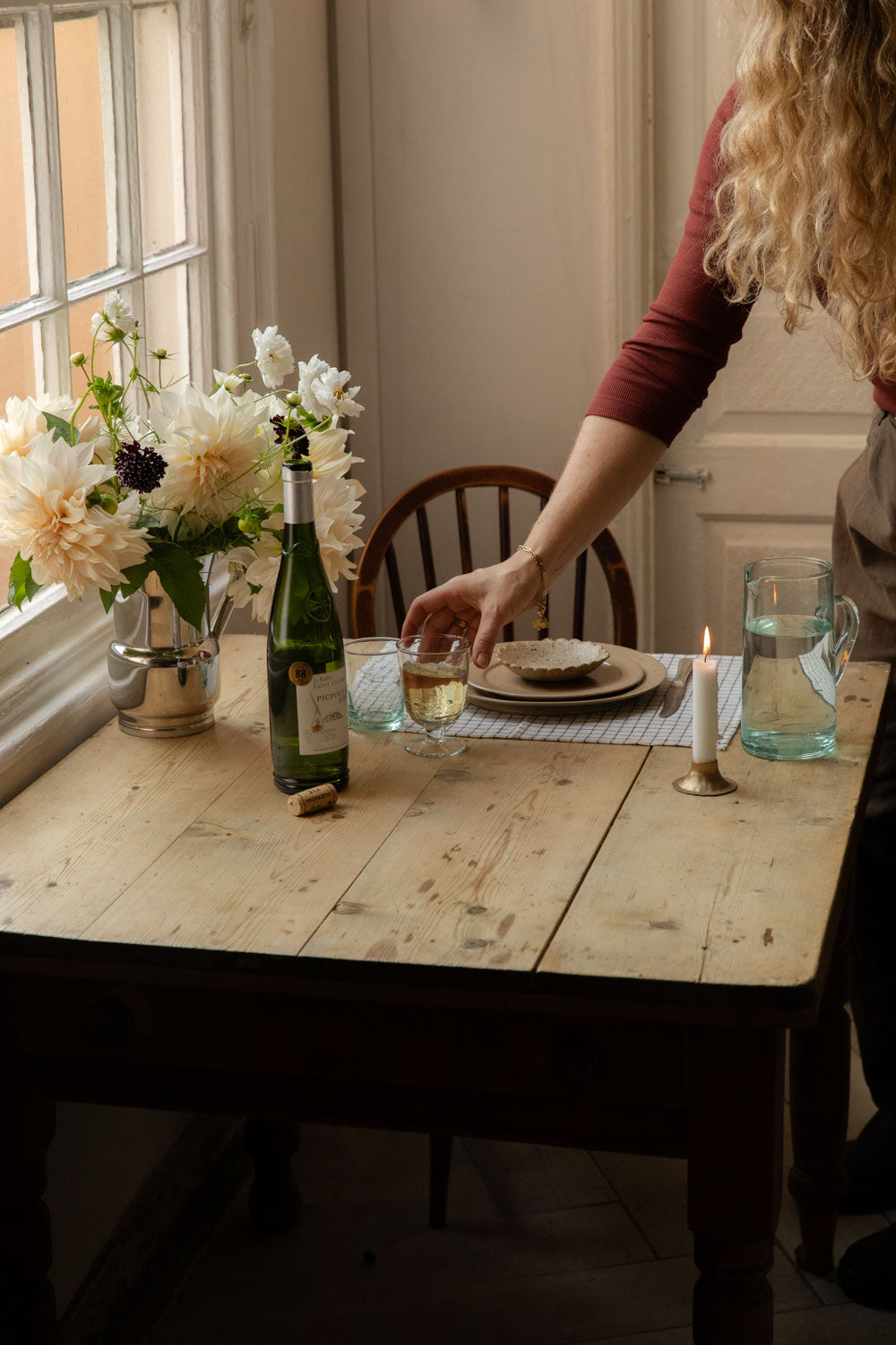 Table for one with wine, a lit candle, dahlias and recycled glass jug by the window