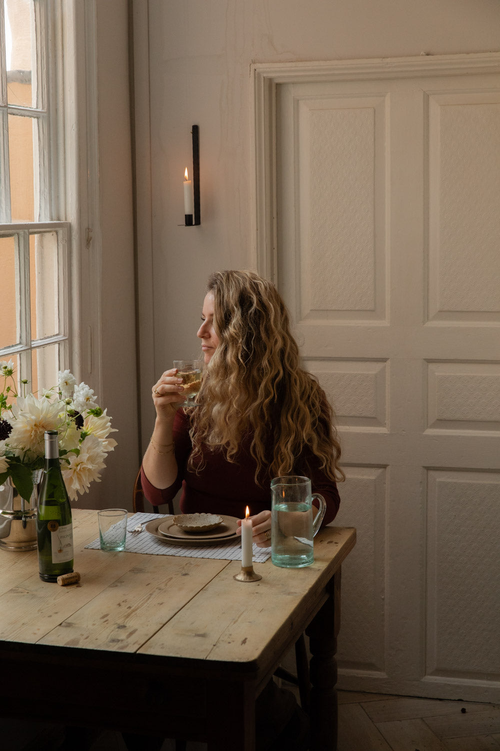 Woman sipping wine at a candlelit table with dahlias and a recycled glass jug