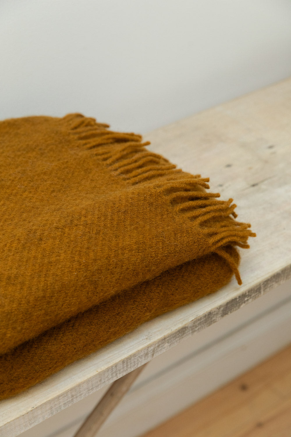 Close-up of Caramel Gotland wool blanket in mustard gold showing soft texture and knotted fringe edge detail on a rustic pale wood table.