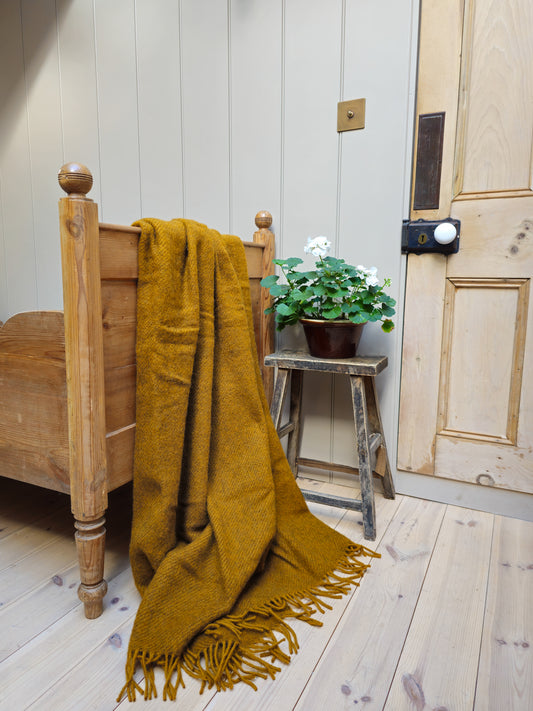 A caramel Gotland wool blanket with fringe draped over the end of a wooden bed frame, with a small potted plant beside it.