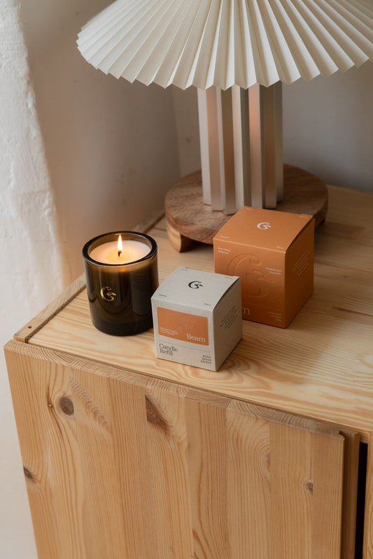 Lit Cedar Beam candle in brown glass with its orange gift boxes under a pleated table lamp
