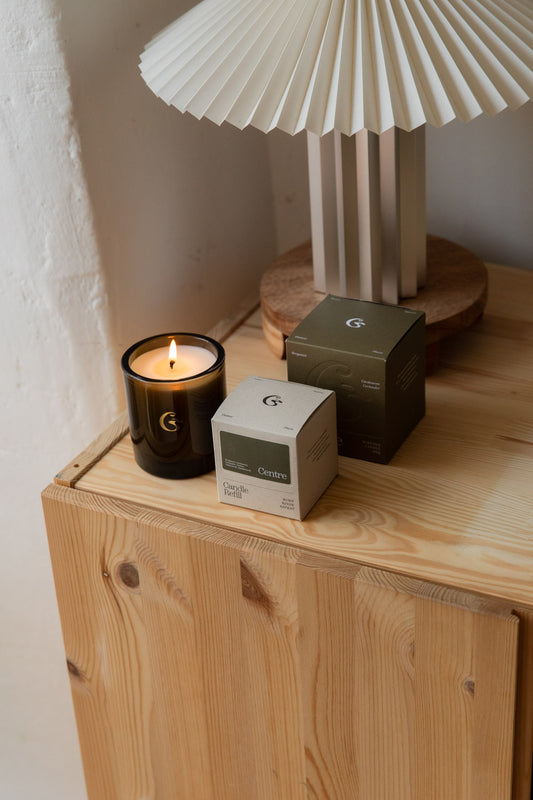 Cedar Centre candle burning in brown glass beside its grey-green and white boxes on a pine cabinet