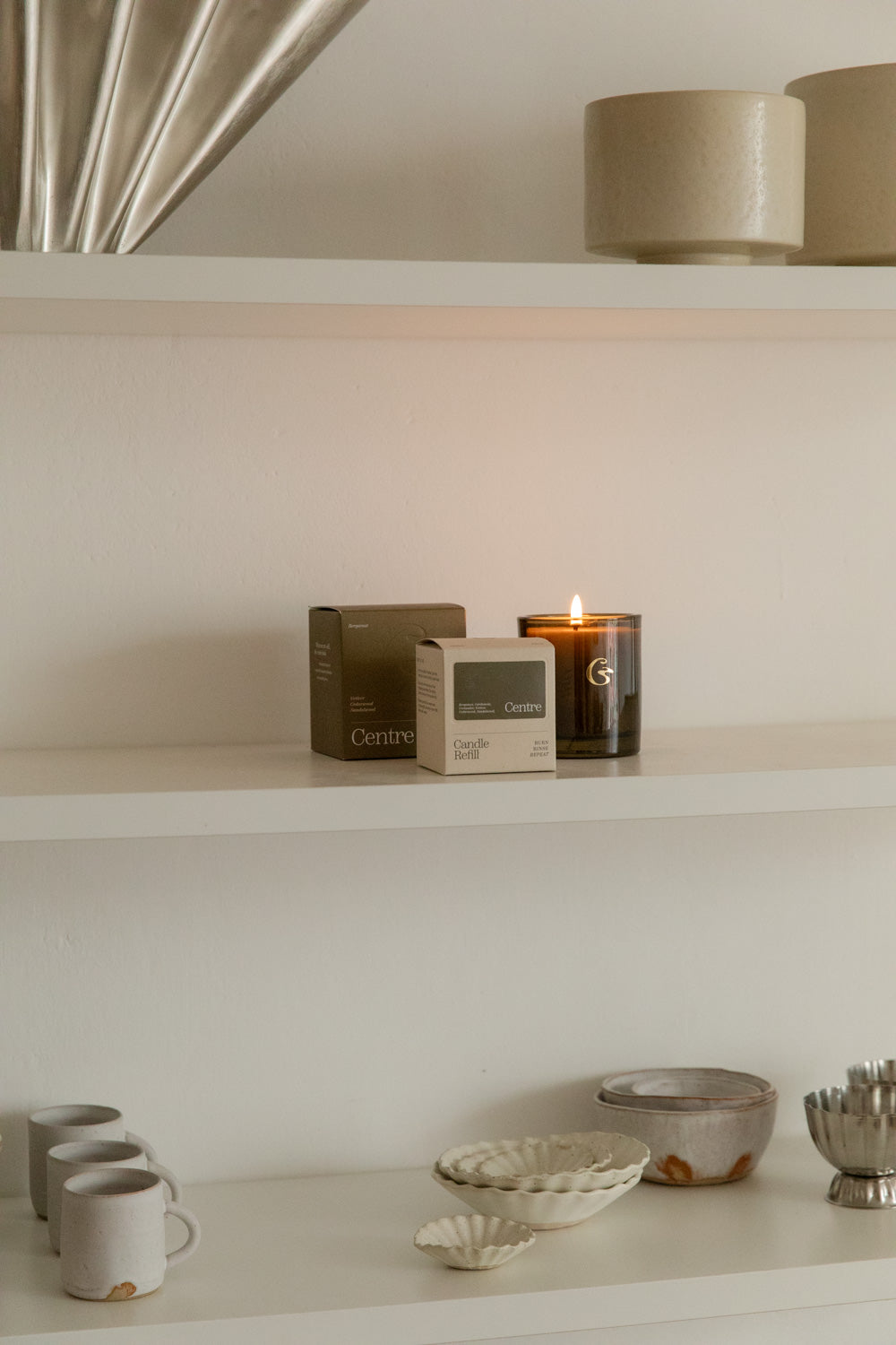 Lit Cedar Centre candle in dark green glass with its boxes on a white shelf above handmade ceramics