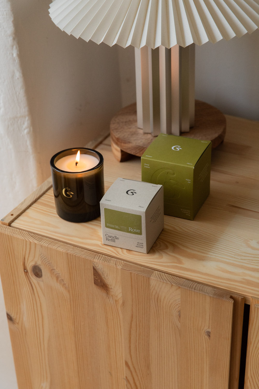 Lit Cedar Rove candle in dark green glass with its olive green gift boxes on a pine cabinet