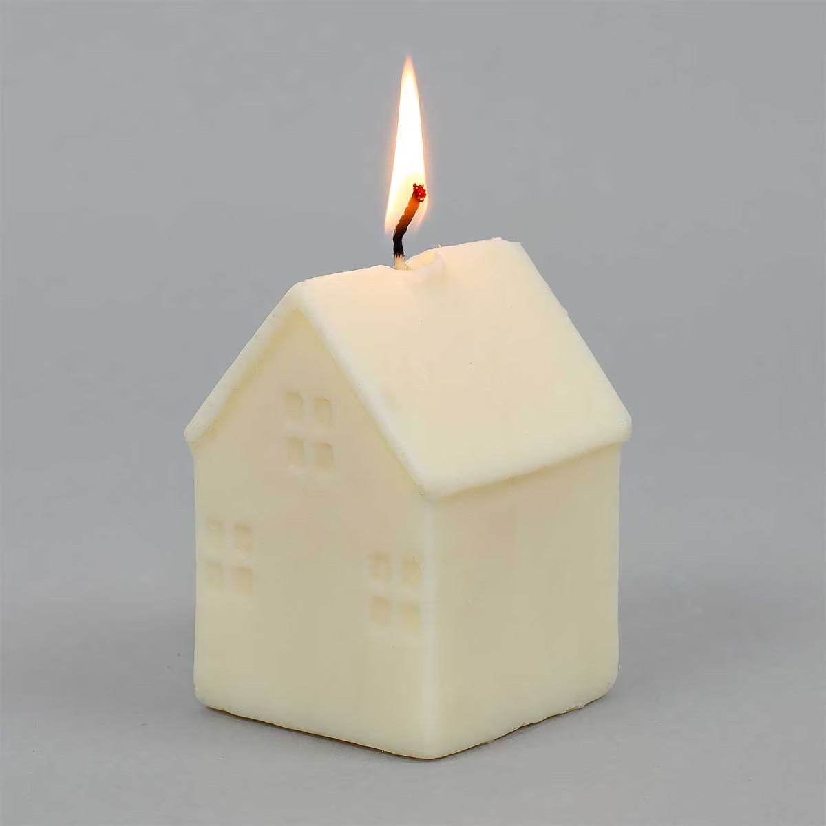A house-shaped candle in a warm cream or off-white color, shown lit with a small flame against a plain light gray background.