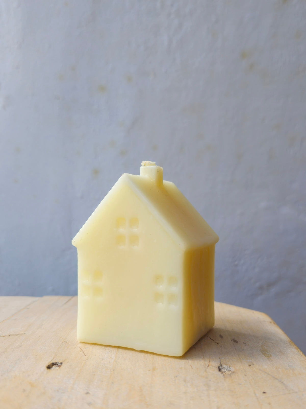 A small house-shaped candle in a pale cream yellow, standing on a light wooden surface against a textured light gray wall.