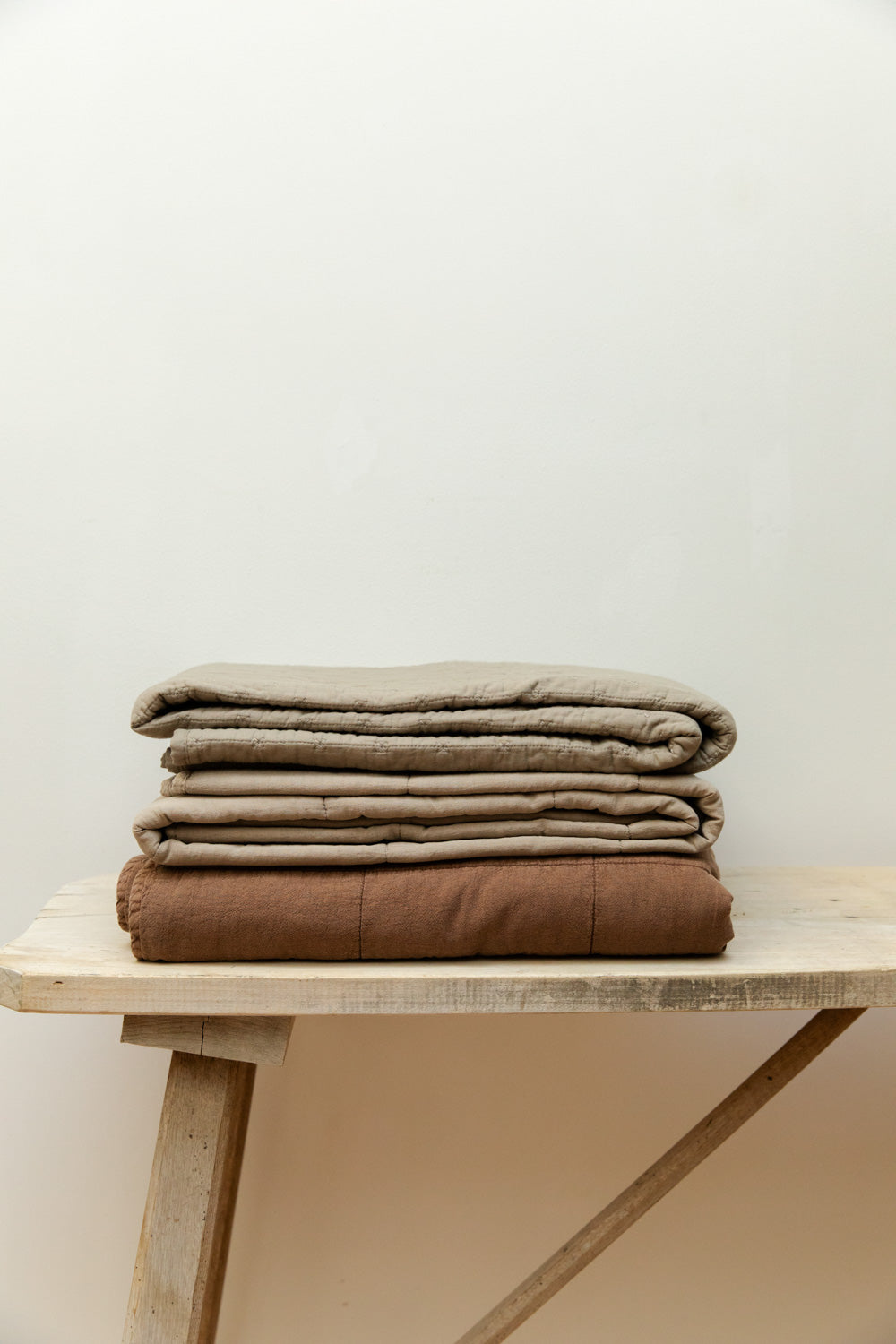 Cotton cross stitch quilts in vintage linen and camel colourways folded and stacked on a pale wood trestle bench against a white wall.