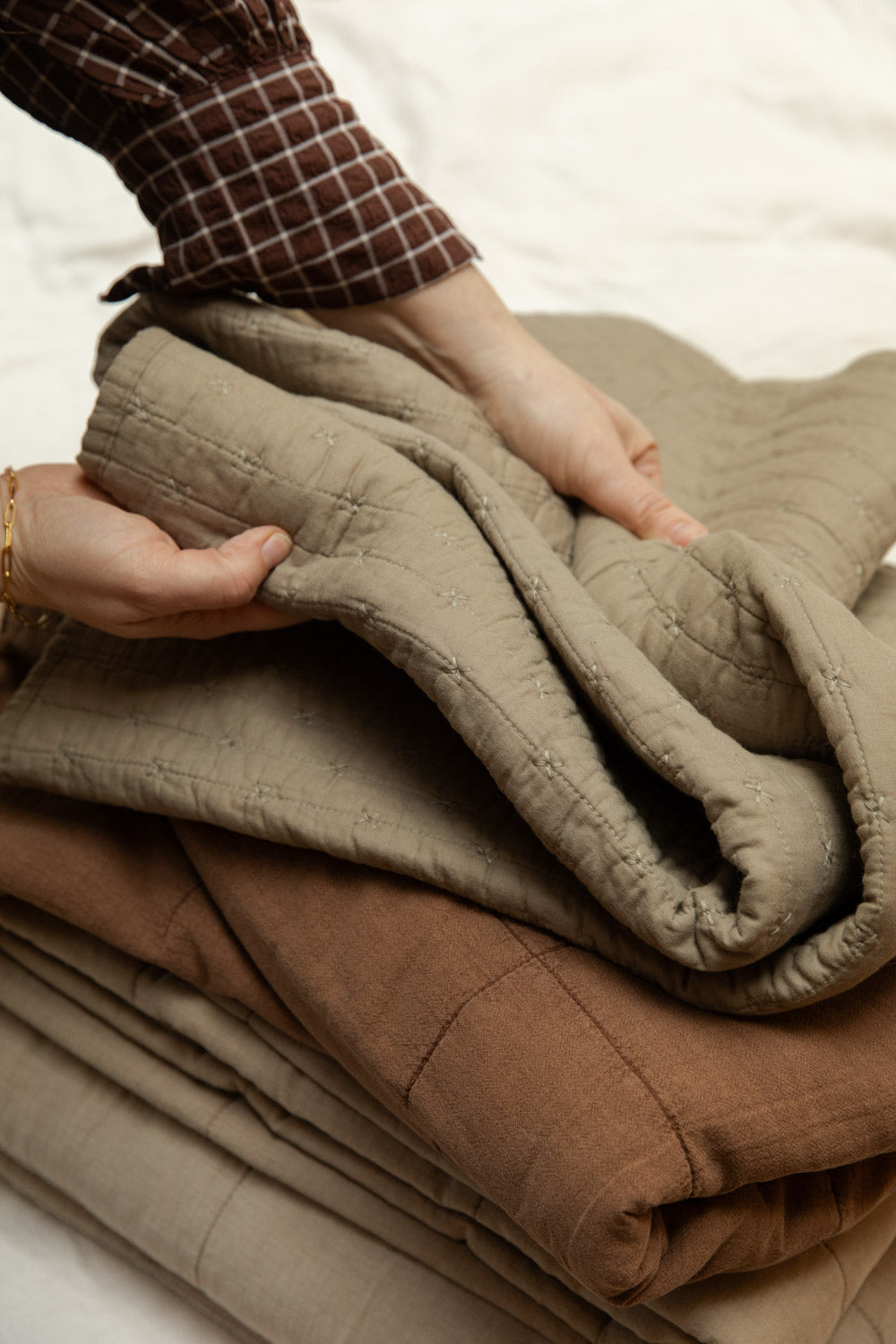 Hands folding a cotton cross stitch quilt in vintage linen on a stack with camel and sand colourways, showing stitched grid texture.