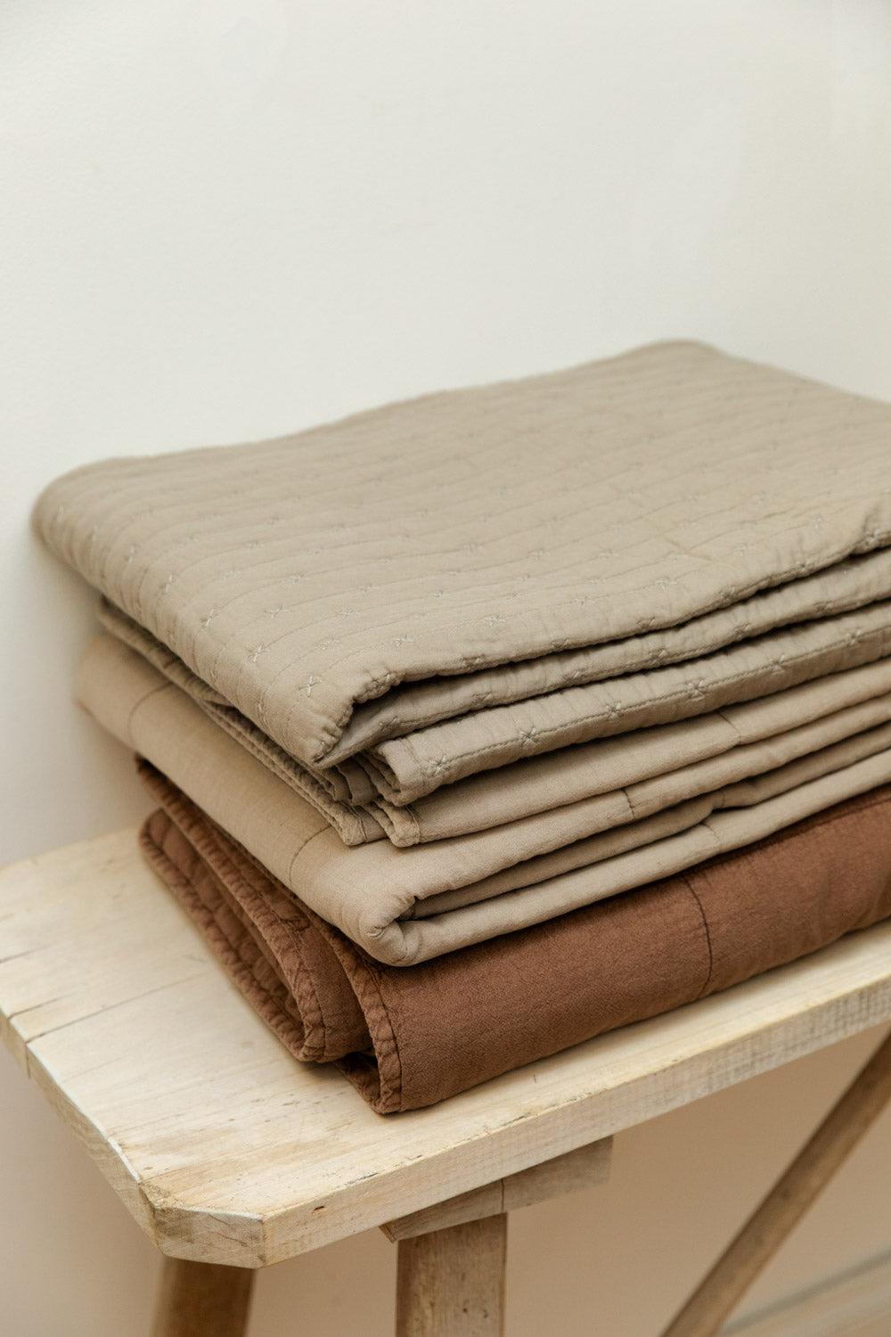 Close view of cotton cross stitch quilts in vintage linen and camel colourways stacked on a pale wood trestle bench, showing quilted edge detail.