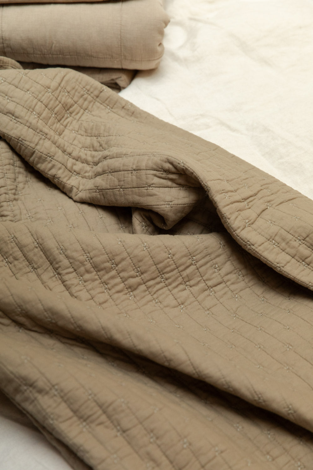 Cotton cross stitch quilt in vintage linen loosely draped on a white linen bed, showing quilted texture and soft crinkled finish.