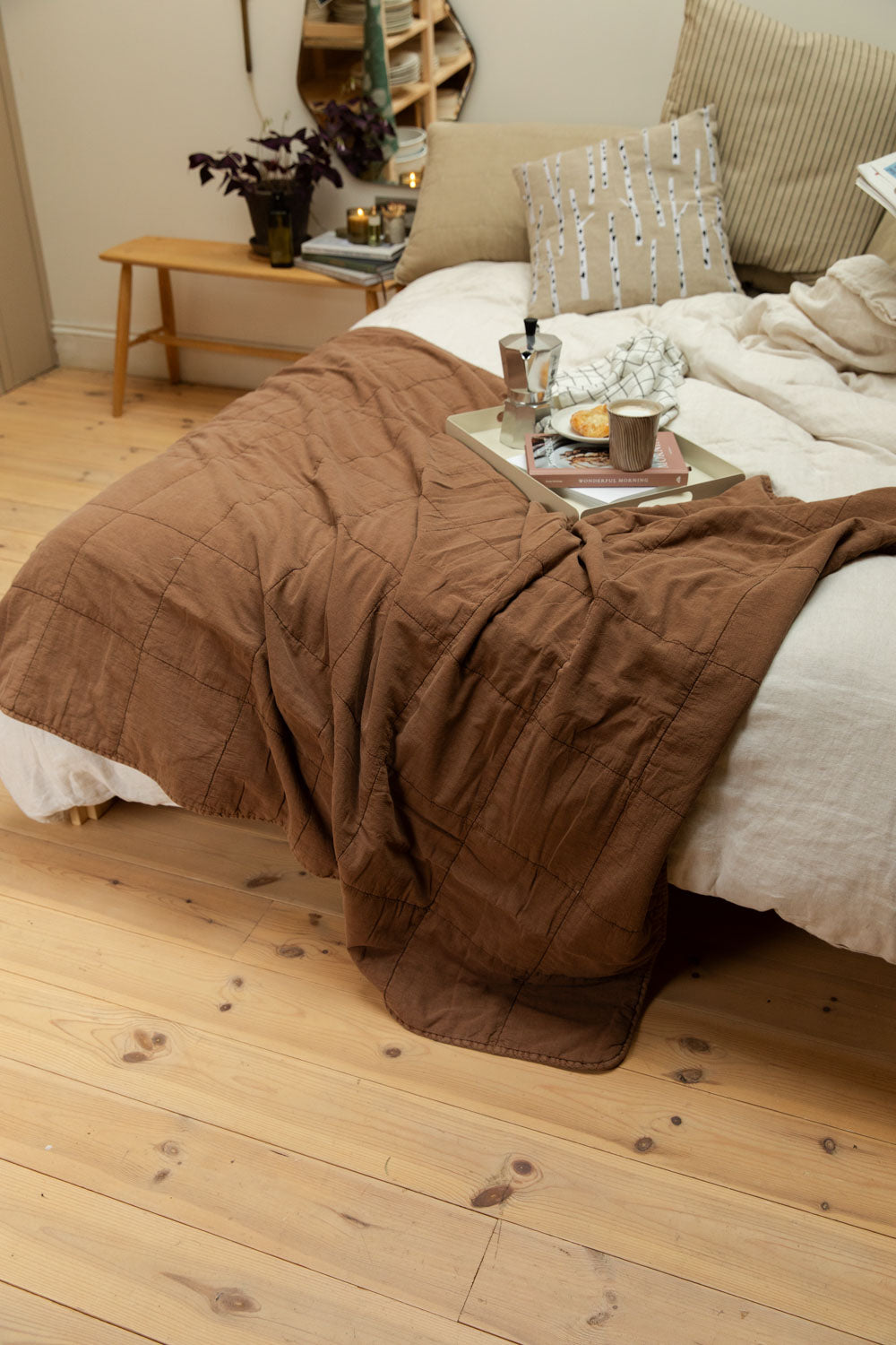 Camel cotton quilt draped over a white linen bed with Bricka breakfast tray, Diva stripe mug, birch tree cushion and candles on pine bedside bench.