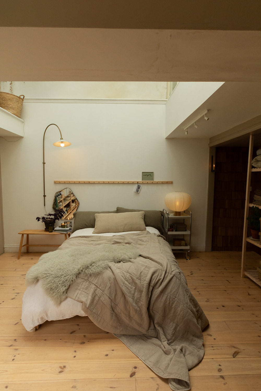 Full bedroom lifestyle shot with cotton quilt in vintage linen draped on bed, sheepskin throw, Blomus Kami lamp and brass floor lamp under skylight.