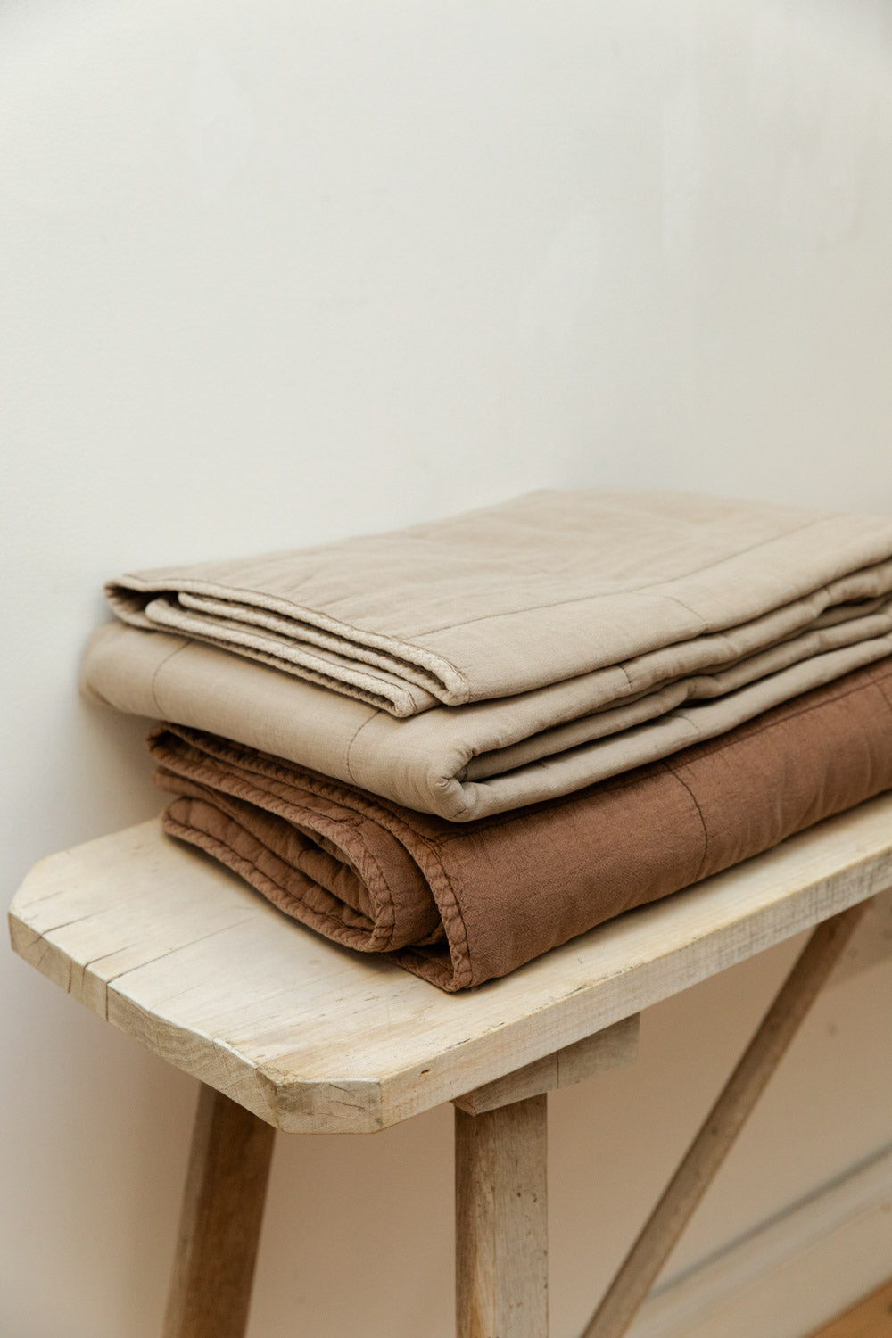 Cotton quilts in vintage linen and rust colourways folded and stacked on a pale wood trestle bench against a white wall.