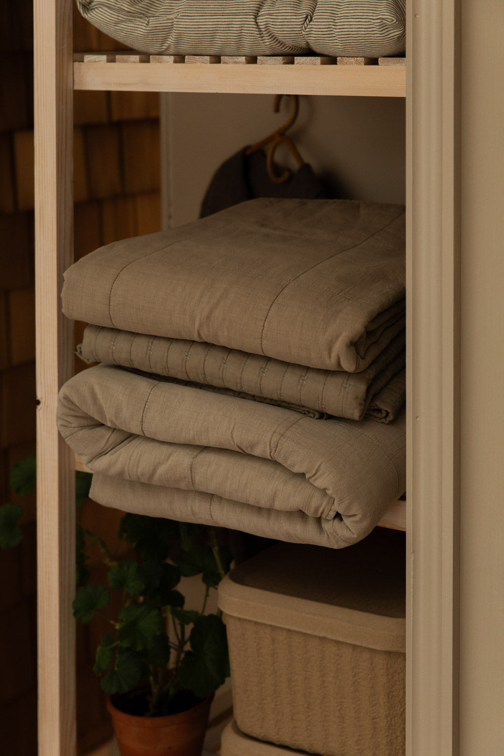 Three cotton quilts in vintage linen folded and stacked on open pine shelving, with rattan basket and potted plant below.