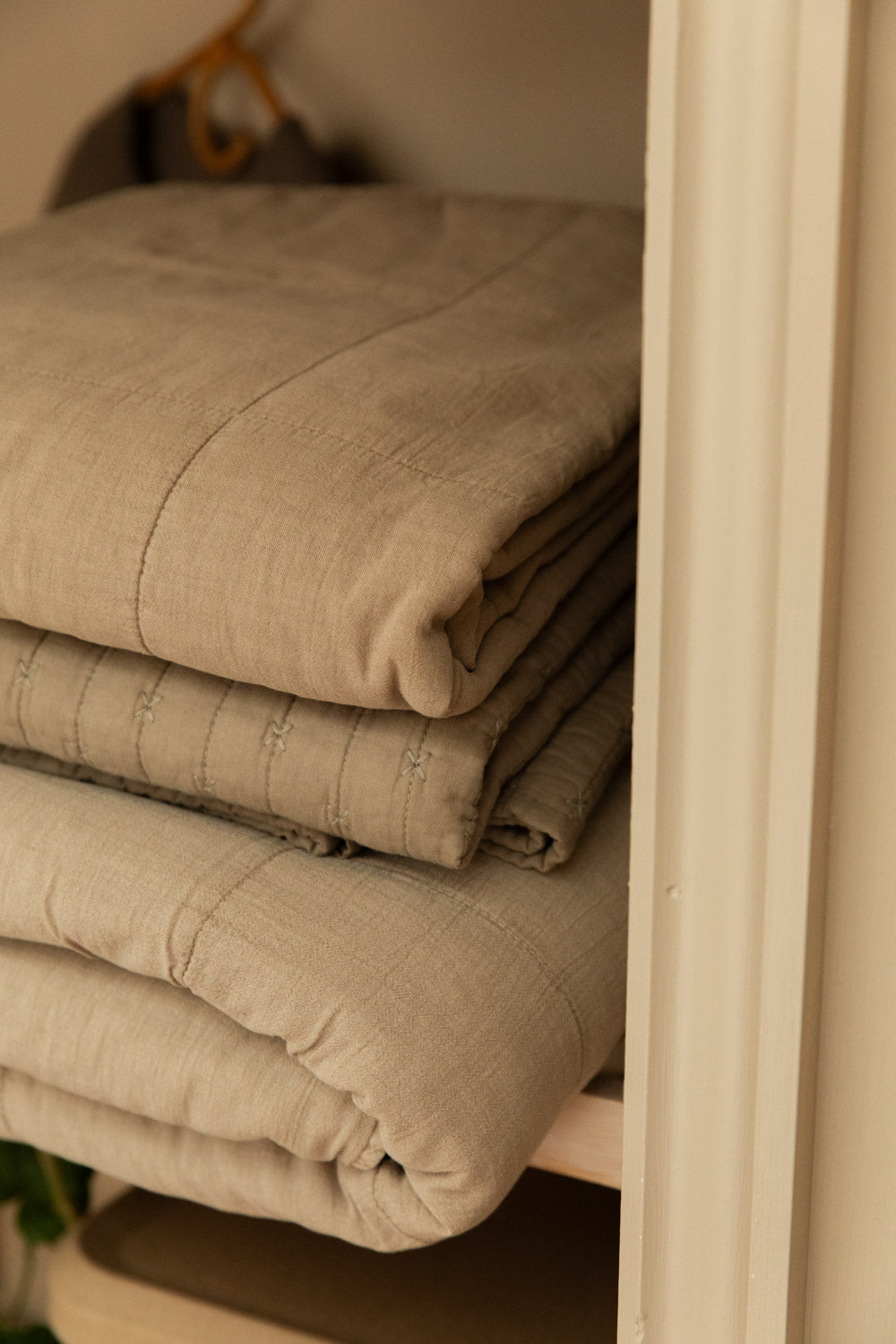 Close detail of cotton quilts in vintage linen neatly folded and stacked on a pine shelf, showing quilted stitch texture and tonal colour.