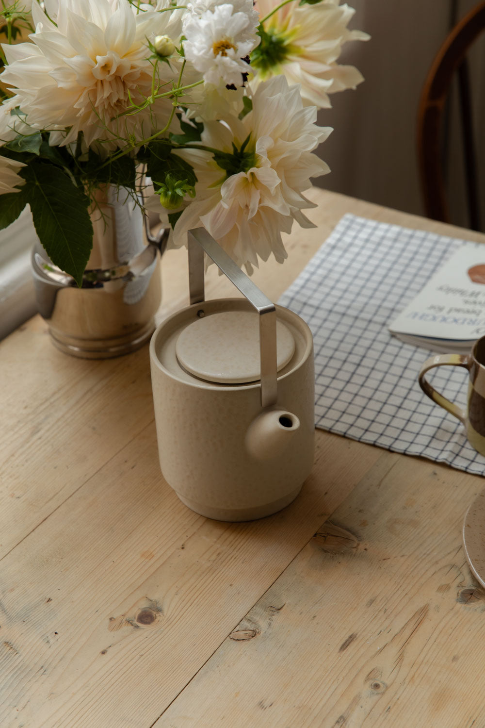 Cream speckled stoneware teapot with a cane handle on a pine table beside a jug of dahlias