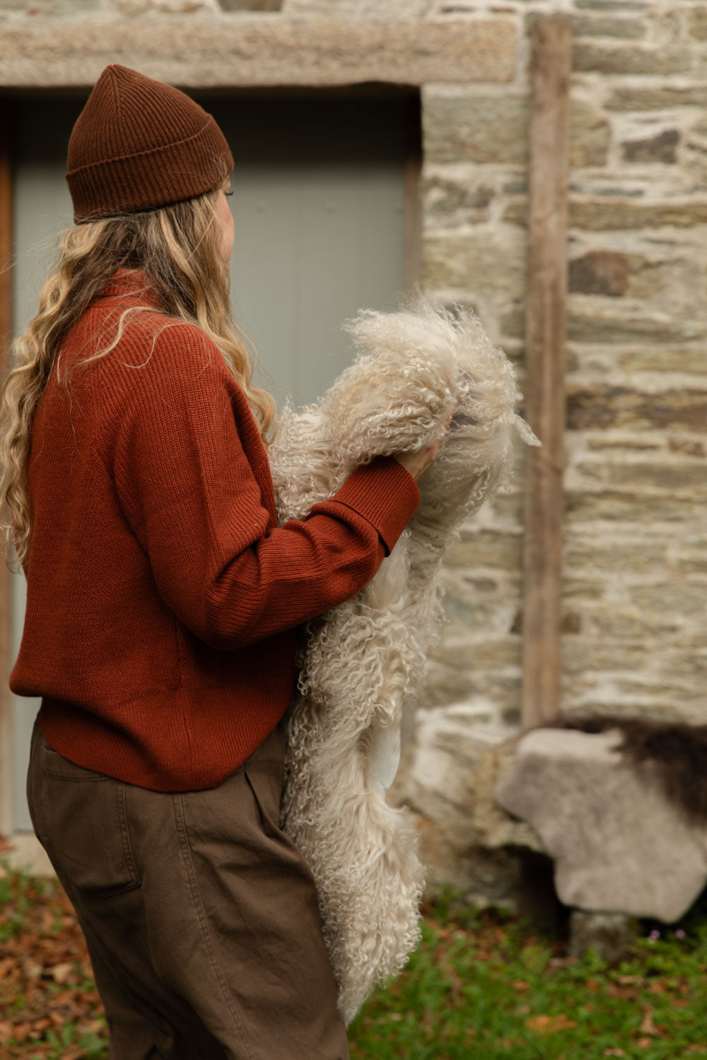 Woman in a rust jumper and brown ribbed beanie holding a long, curly cream Tibetan sheepskin rug against an old stone wall
