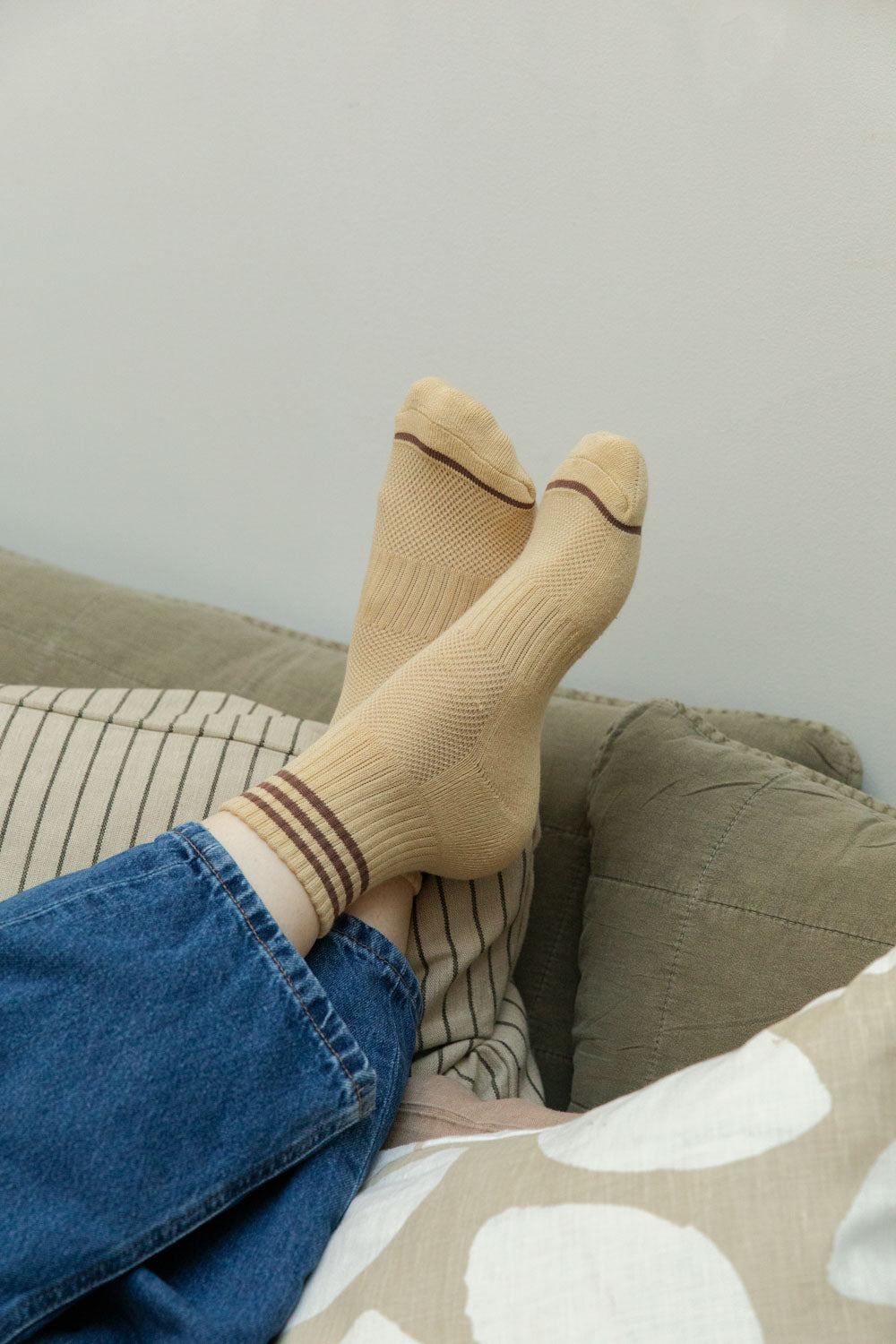 Feet raised wearing daisy yellow textured sports socks with burgundy stripe cuffs, denim jeans, resting on striped and olive cushions