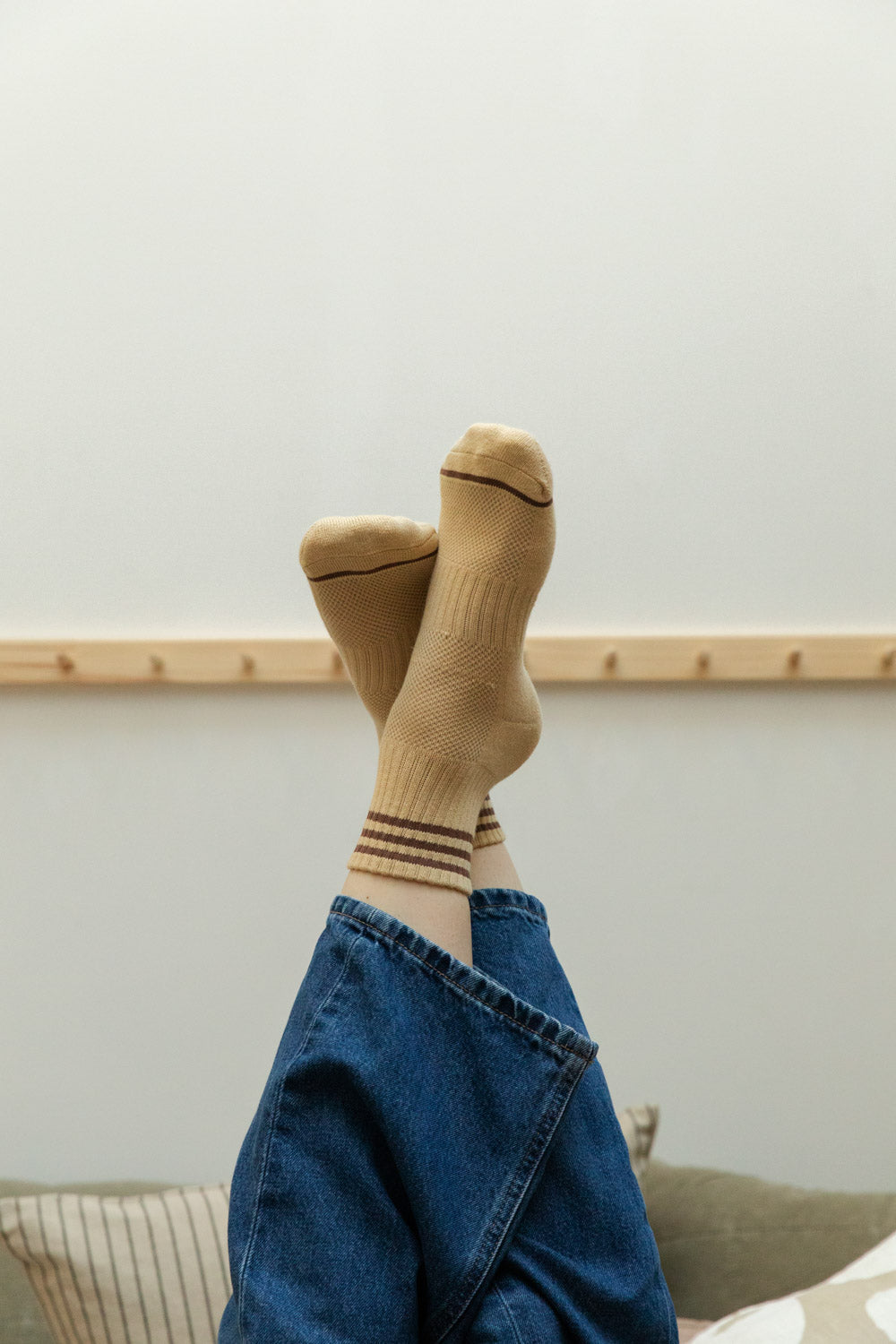 Legs raised in denim jeans wearing sand yellow textured sports socks with burgundy stripes against a soft grey-green wall and pine peg rail