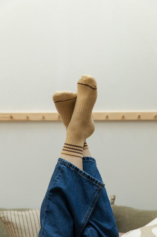 Legs raised in denim jeans wearing sand yellow textured sports socks with burgundy stripes against a soft grey-green wall and pine peg rail