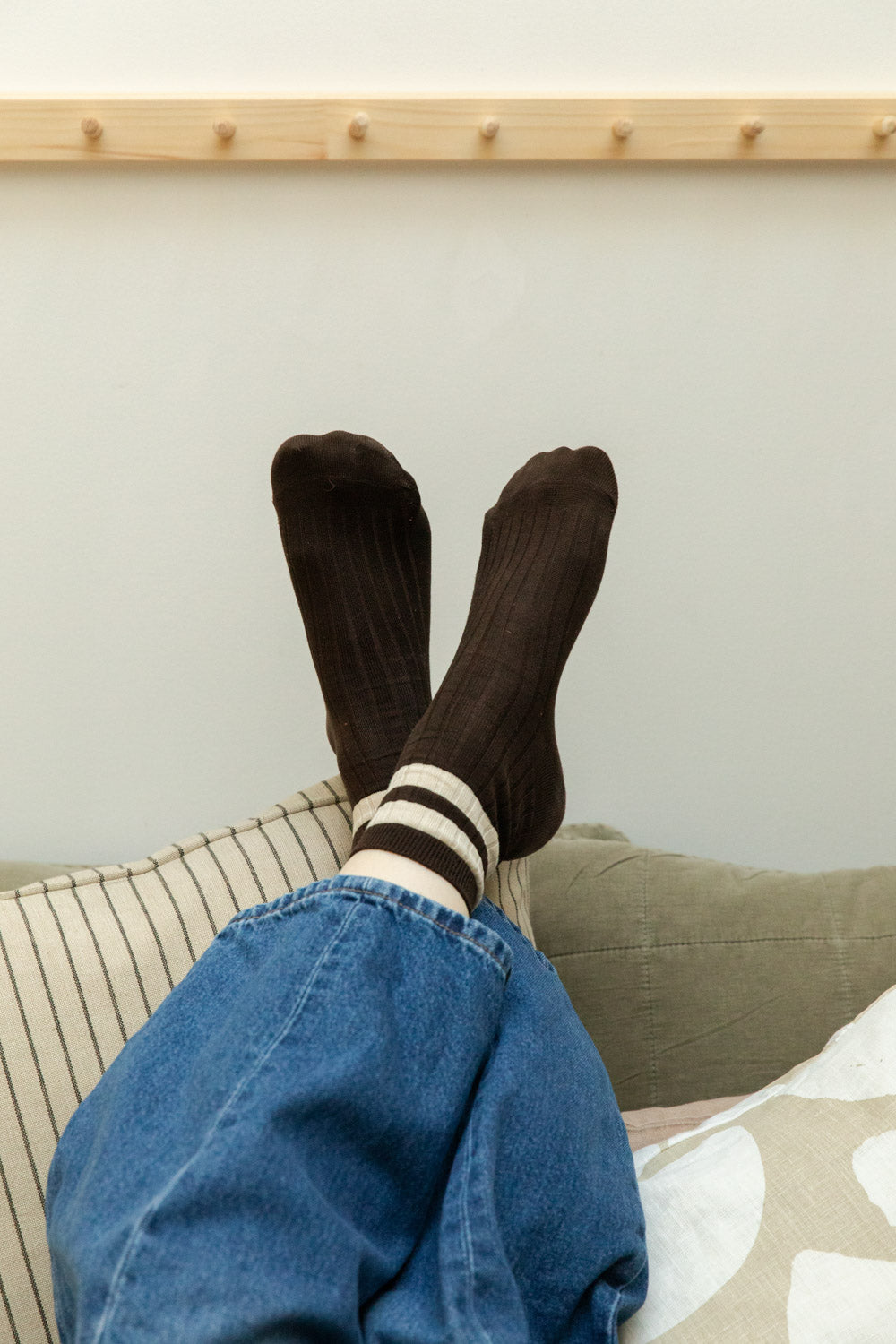 Feet raised wearing dark brown ribbed socks with cream stripe cuffs, denim jeans, resting on striped and olive quilted cushions