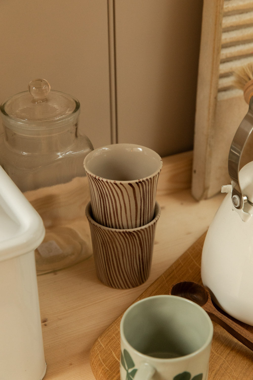 Two stacked Diva mugs with brown stripe pattern on a wooden kitchen shelf, styled with a glass jar and white enamel kettle.