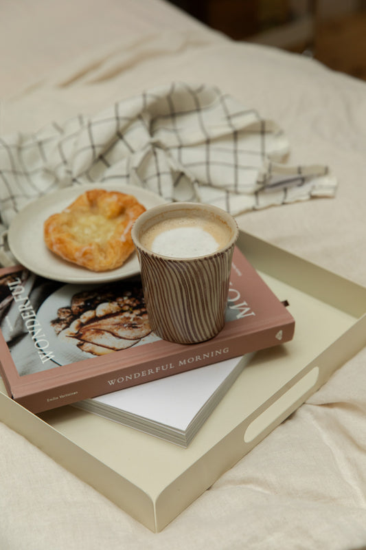 Close detail of Diva stripe mug with frothy coffee on a Bricka off-white tray, with pastry and Wonderful Morning book on white linen bed.