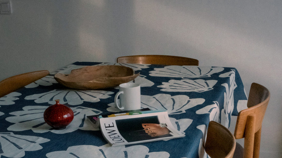 A navy blue tablecloth with a large white shell pattern draped over a wooden dining table, surrounded by wooden chairs in a bright room.