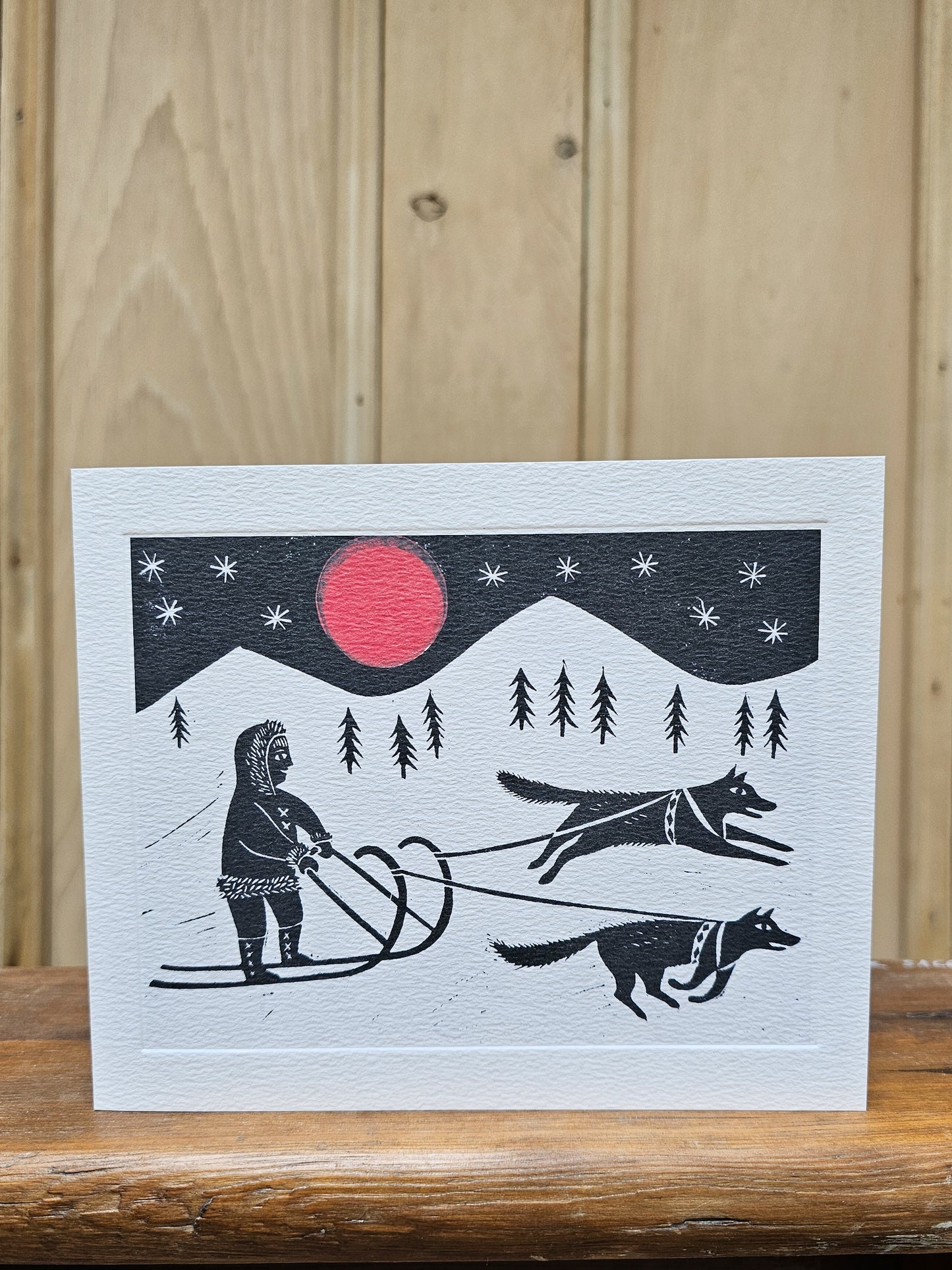 Greeting card reproducing Emily Sutton's linocut print of a sleigh ride, showing a figure in traditional dress being pulled by two huskies under a red moon and starry night sky