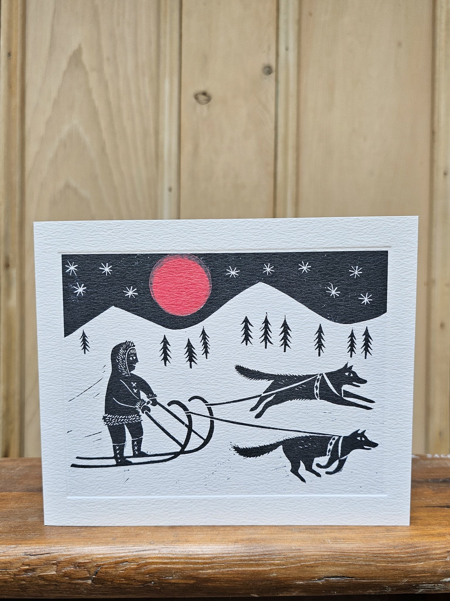 Greeting card reproducing Emily Sutton's linocut print of a sleigh ride, showing a figure in traditional dress being pulled by two huskies under a red moon and starry night sky