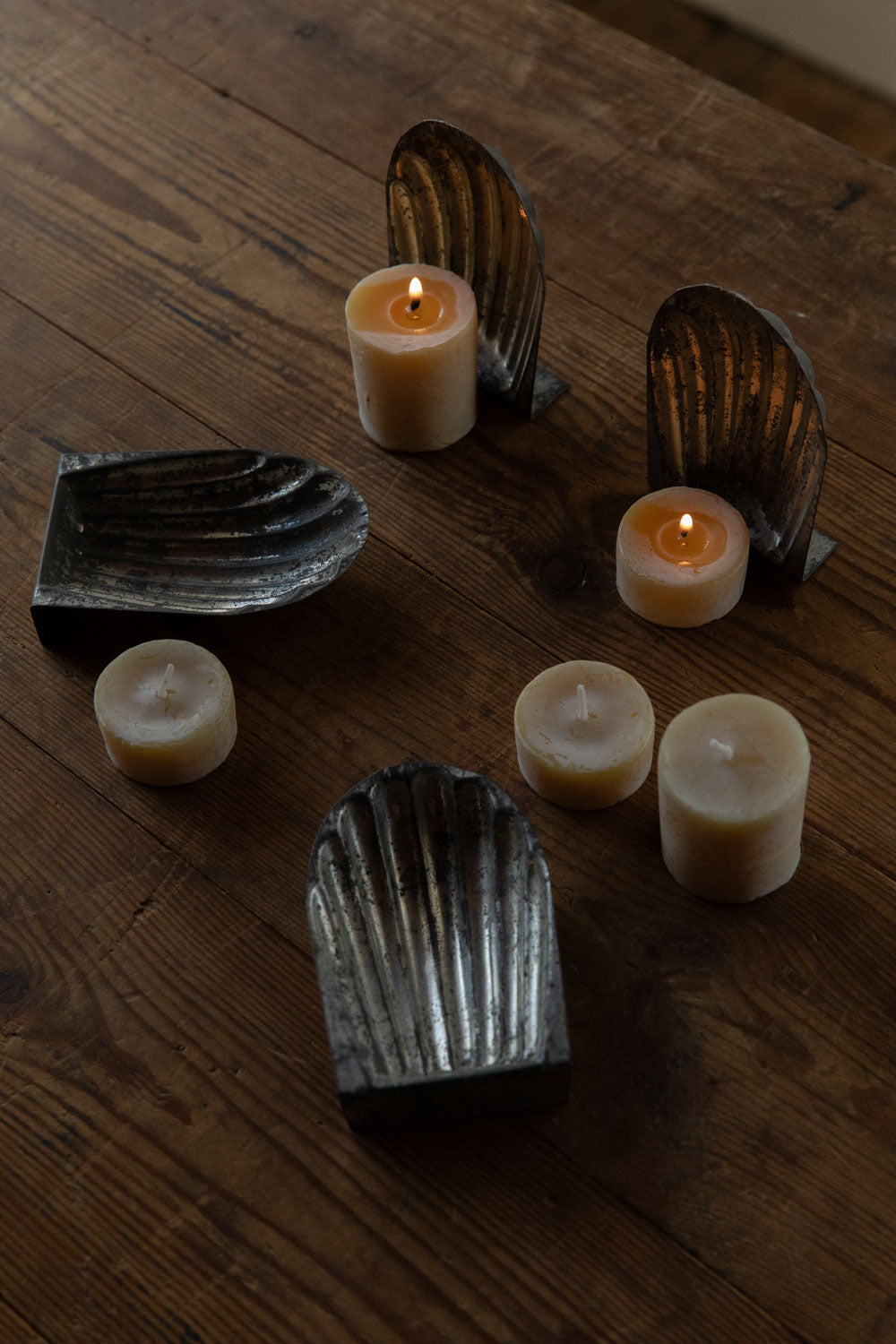 Decorative metal shell moulds shown next to both lit and unlit English beeswax tealight candles on a wooden surface.