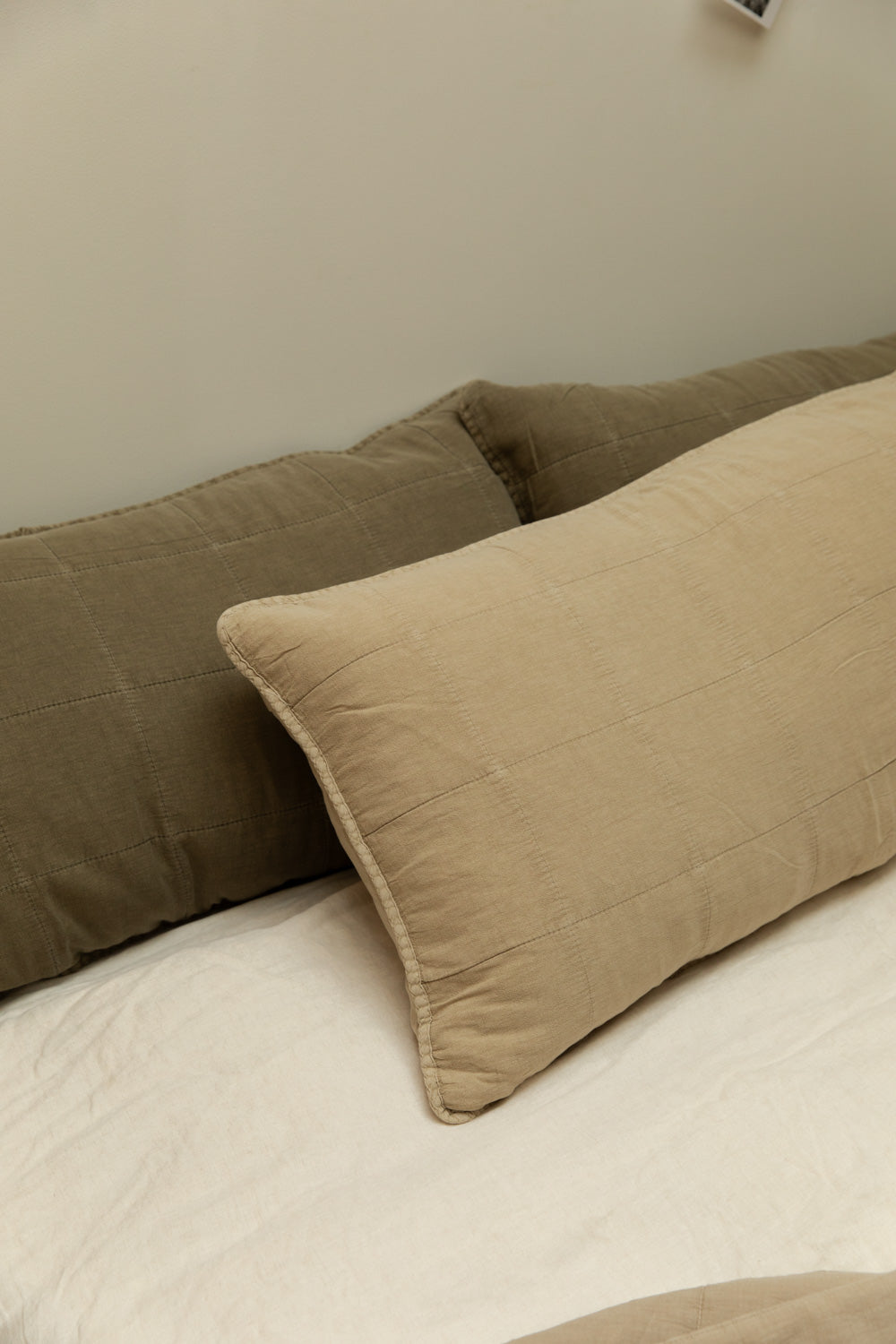 Eucalyptus green and fog quilted rectangular headboard cushions layered on a white linen bed against a soft grey-green wall