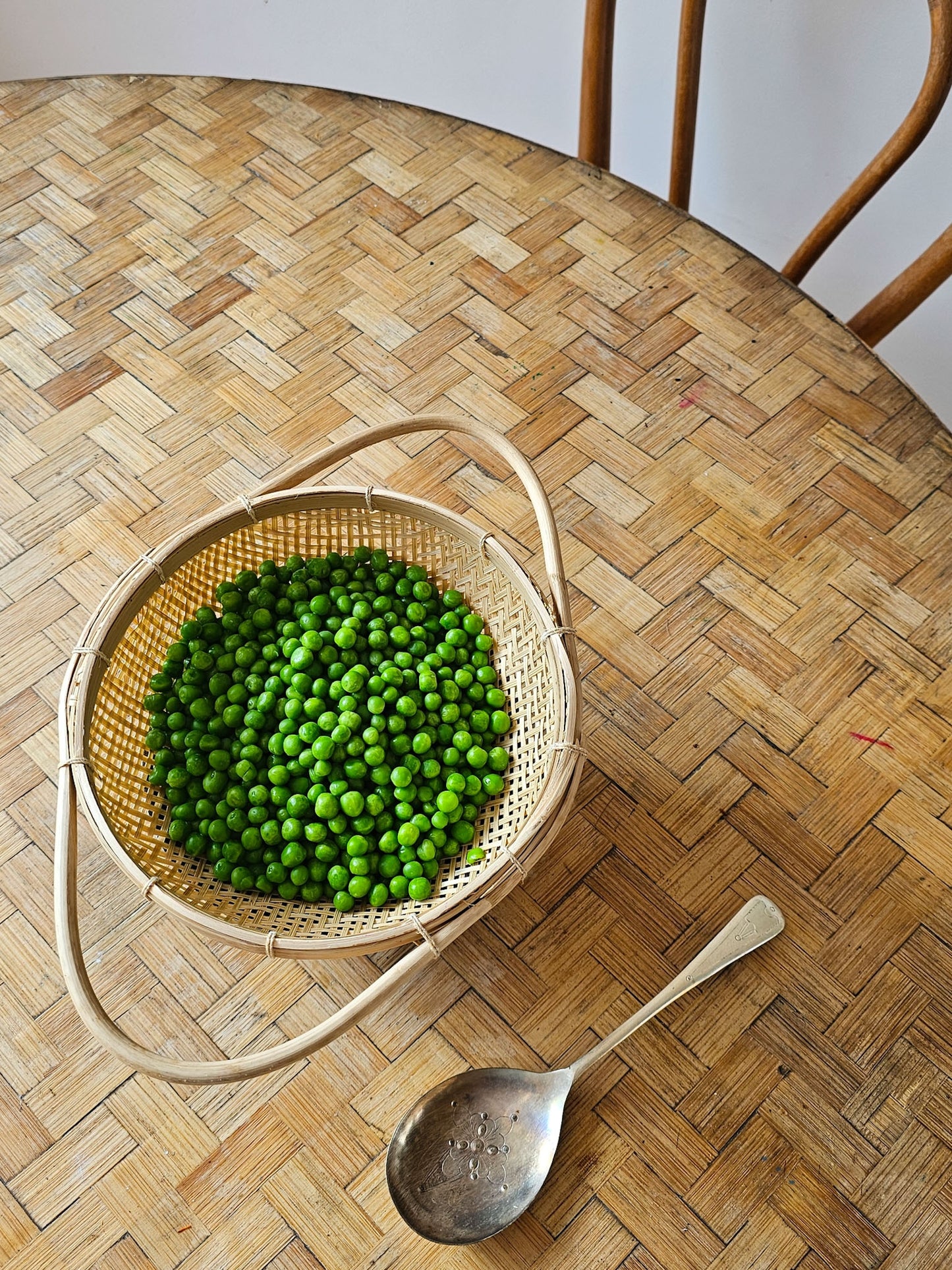 A round woven bamboo strainer filled with bright green peas, placed on a light wood herringbone-patterned table next to a metal spoon.