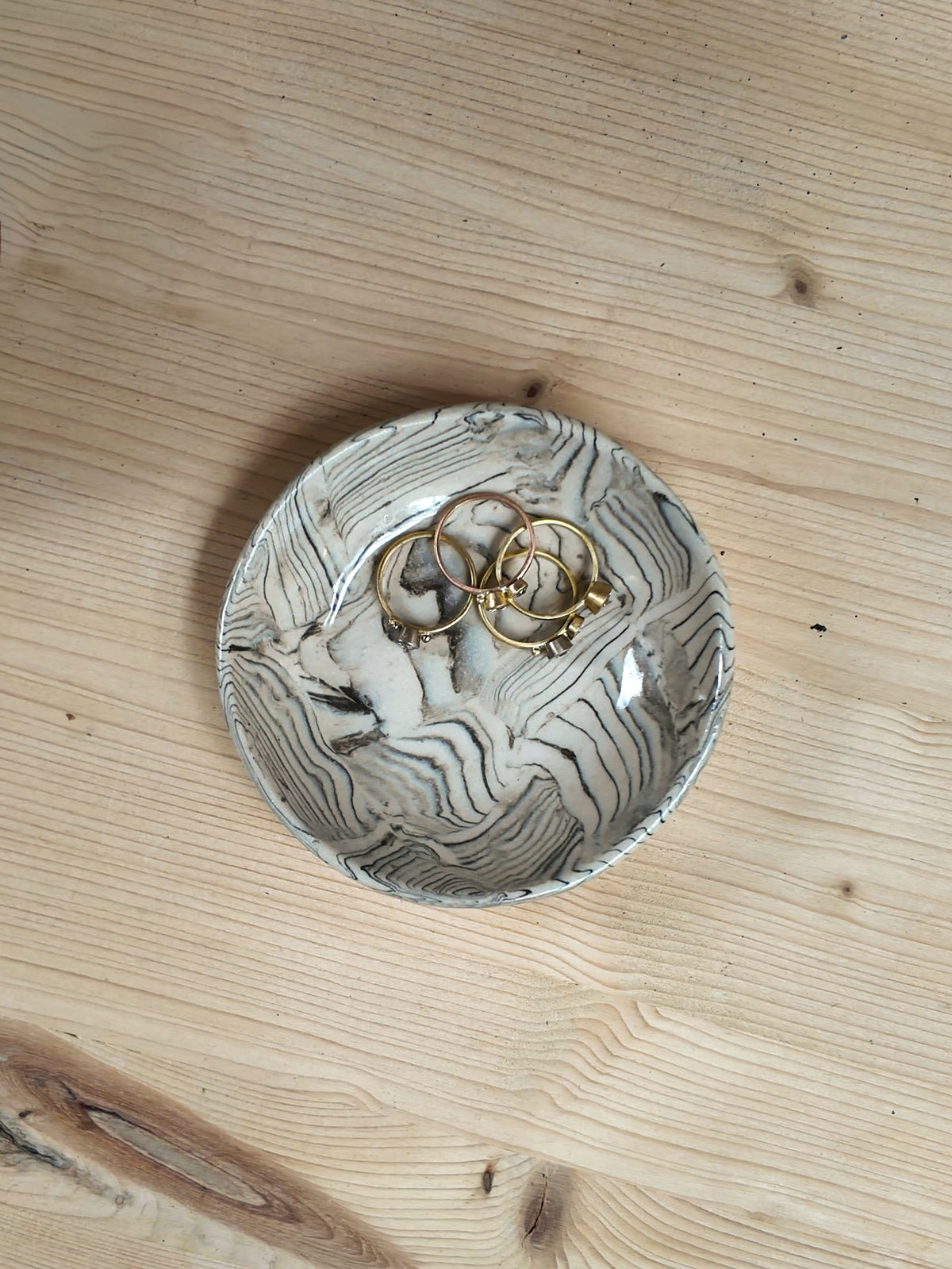 A small round ceramic dish with a gray and white wavy wood-grain pattern, holding three gold rings, placed on a light wood tabletop.