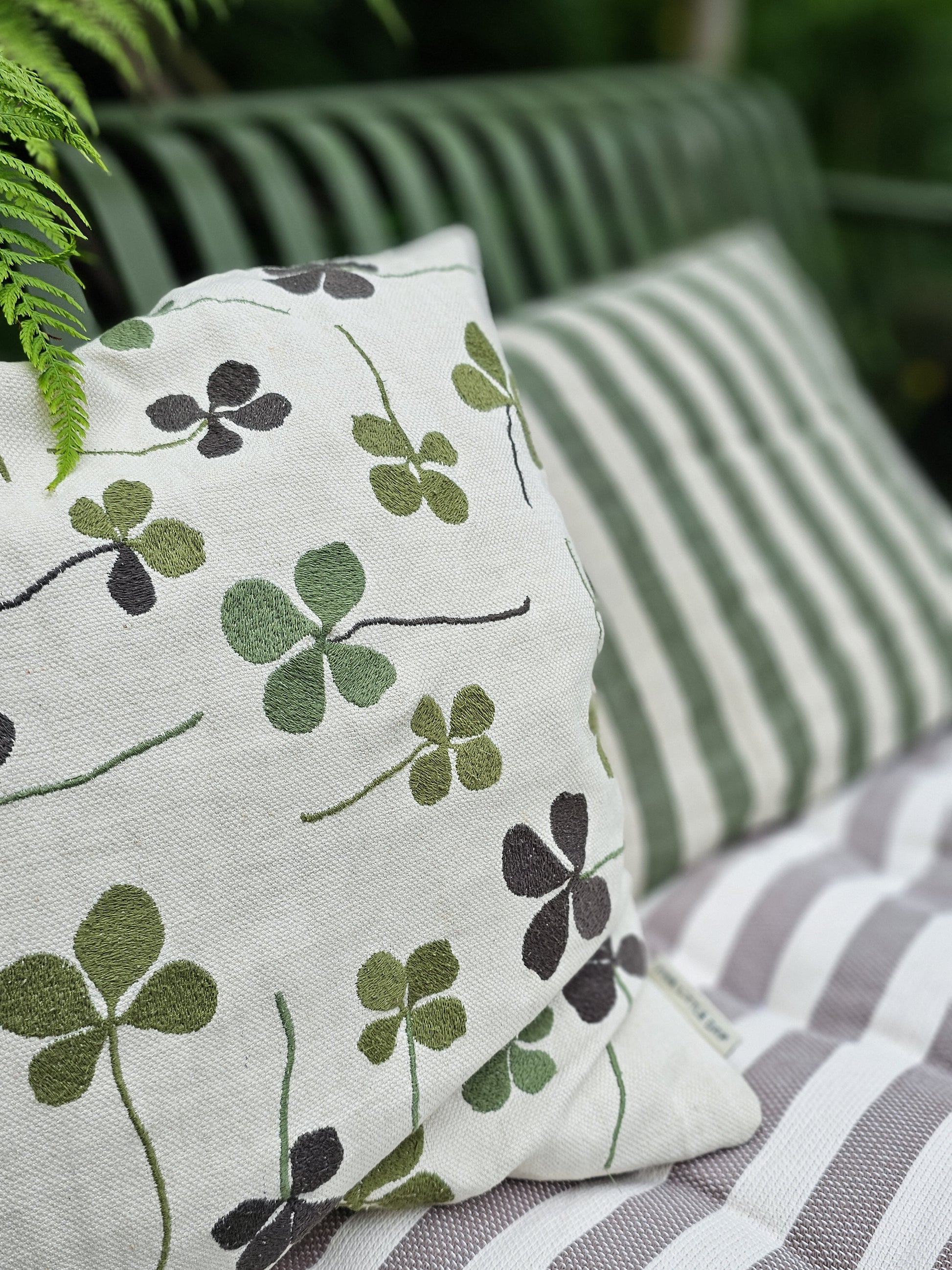 A linen coloured cushion with a green clover embroidery pattern on an outside bench.