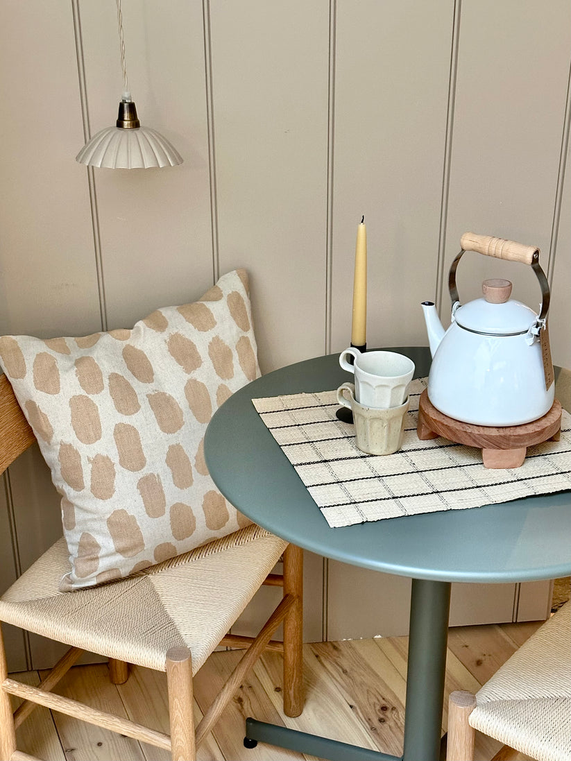 A square-shaped cushion with a beige background and a pattern of sand coloured dots on a wooden chair next to a green metal table with kitchen items on it.