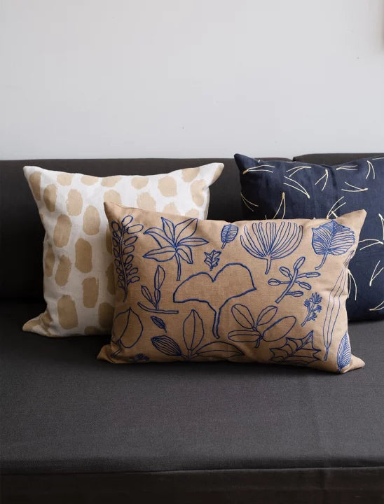 A square-shaped cushion with a beige background and a pattern of sand coloured dots nestled amongst other cushions on a black sofa.