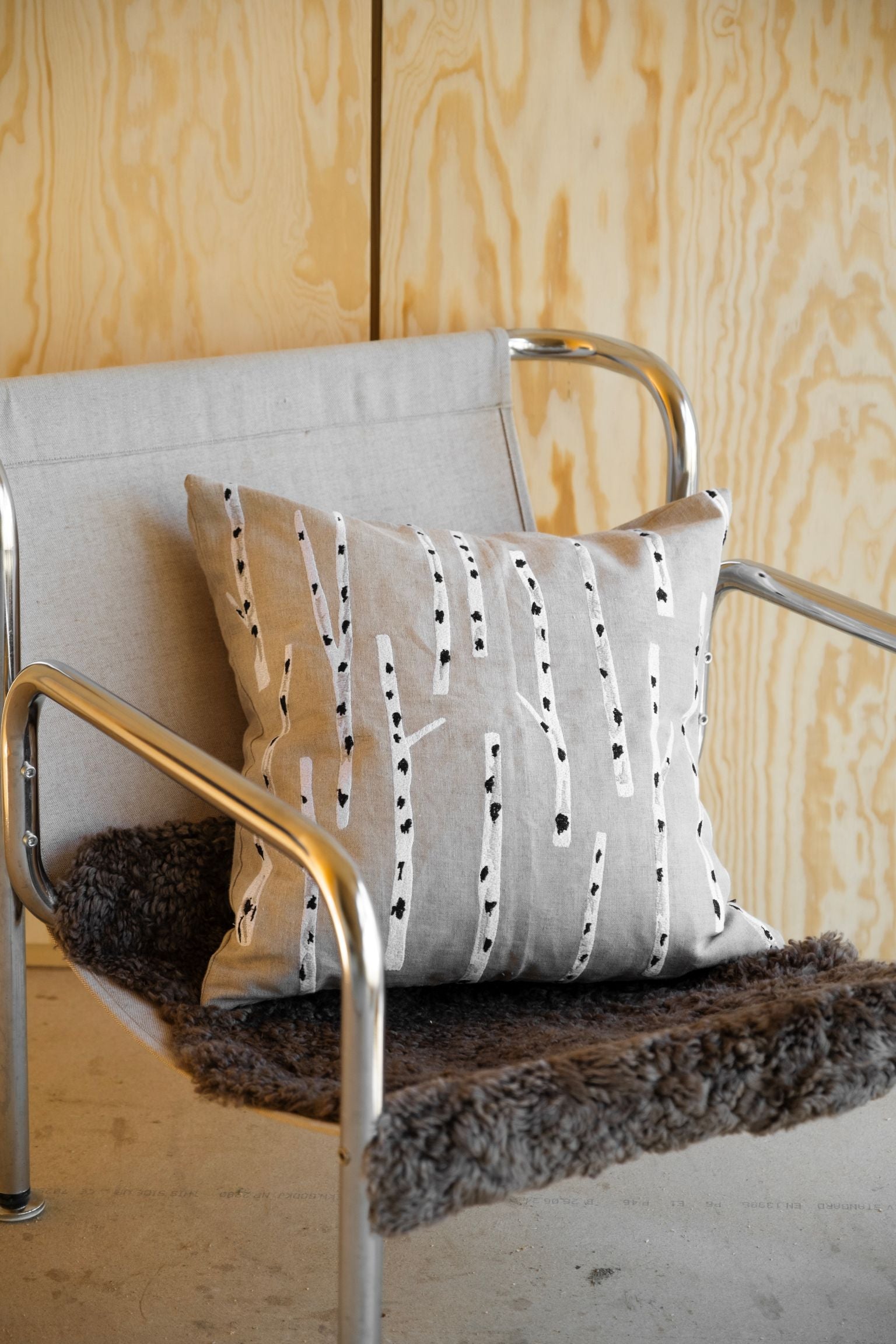 Decorative embroidered Fine Little Day birch pillow sitting on a metal and fabric chair against a wooden wall.