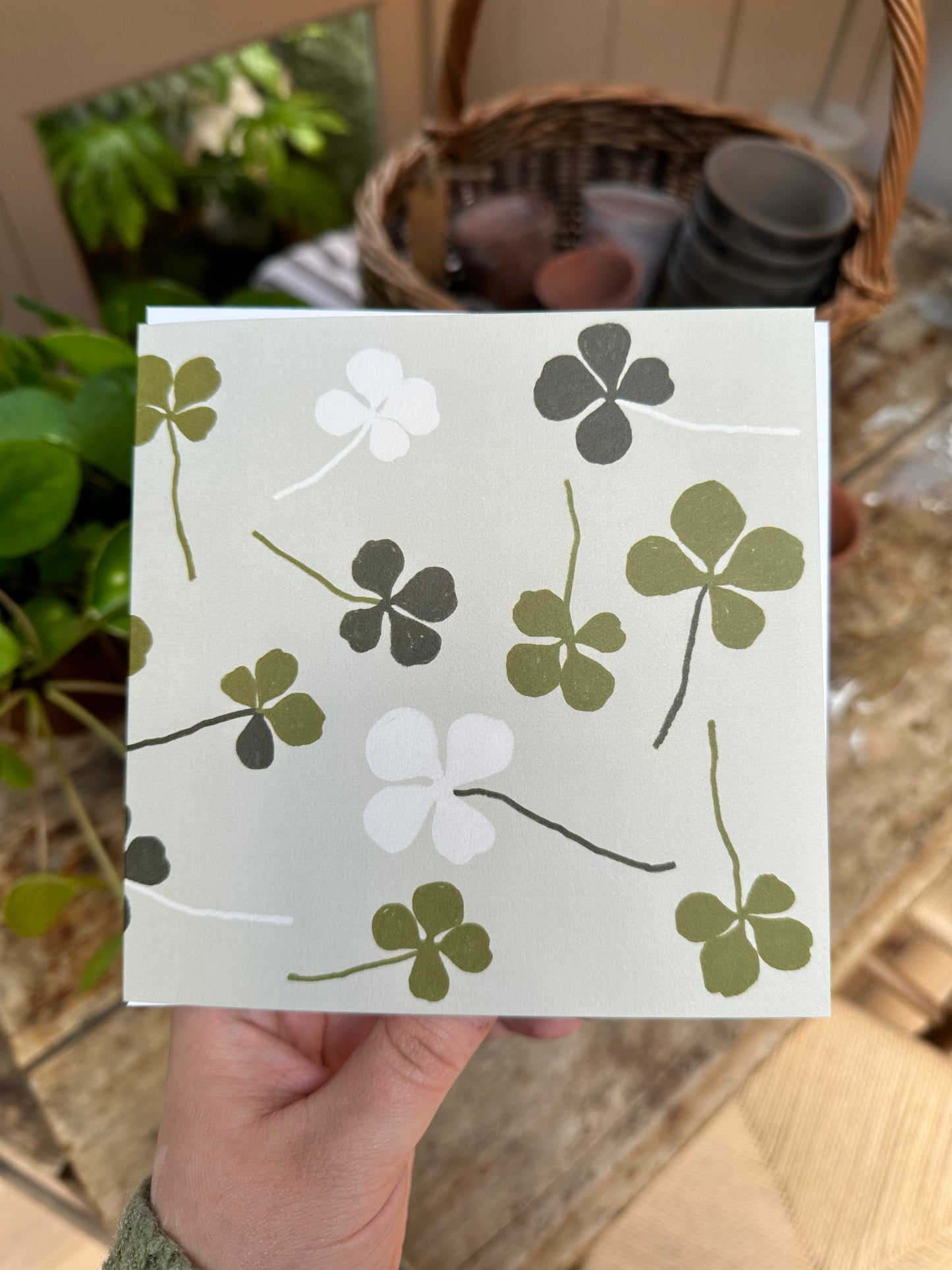 A hand holding a greeting card with a four leaf clover design and green accents on a beige background.