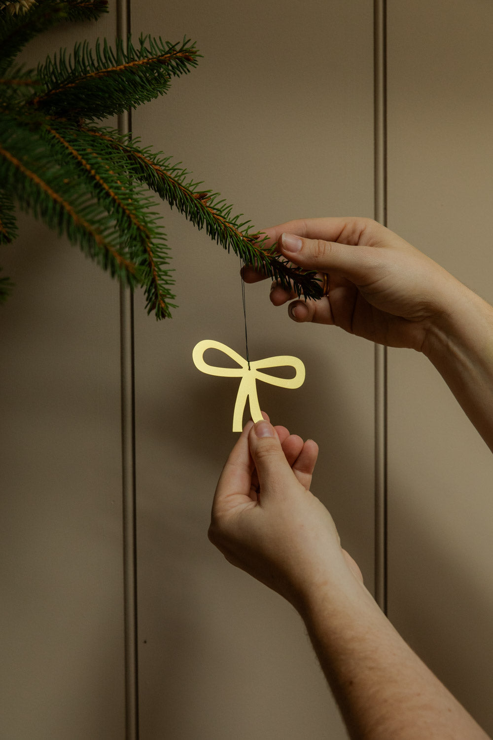 Person hanging a golden bow decoration on Christmas tree branch against a neutral background.
