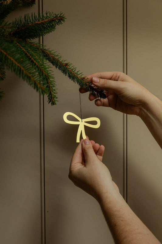 Person hanging a golden bow decoration on Christmas tree branch against a neutral background.