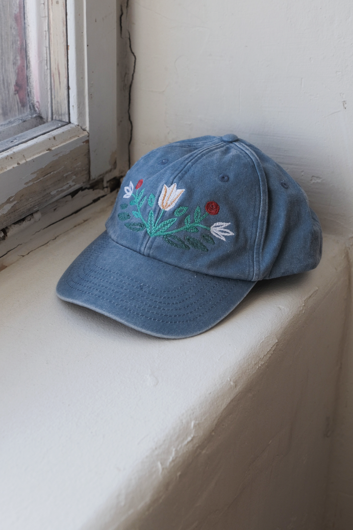 A washed denim cap with a floral kurbits embroidery on the front panel, placed on a window sill with a visible adjustable strap back.