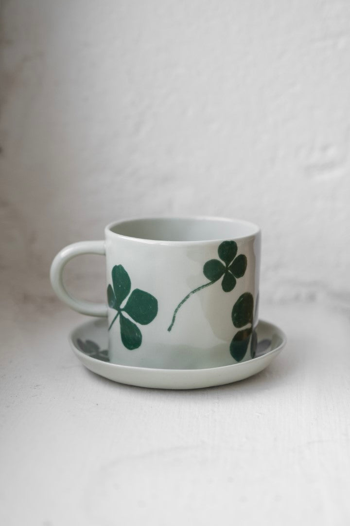 A round porcelain plate and cup with a green four-leaf clover design, placed on a white surface.