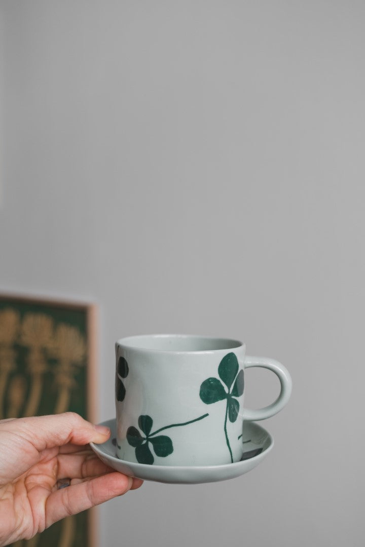 A hand holding a round porcelain plate and cup with a green four-leaf clover design, placed on a white surface.