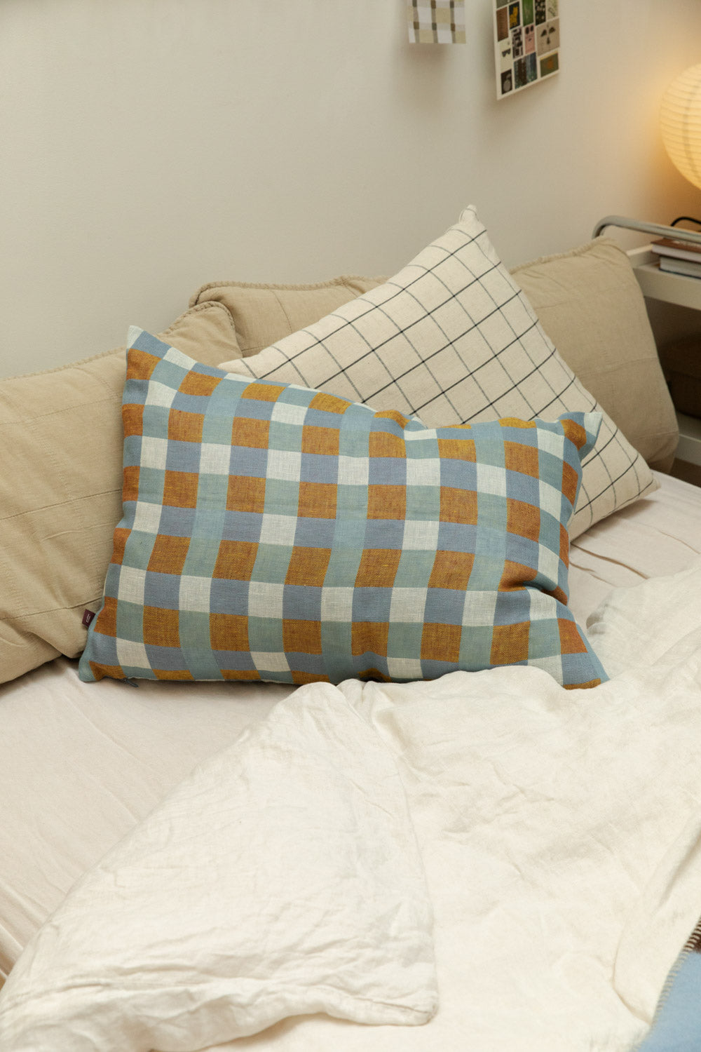 Fine Little Day Rutan linen cushion cover in blue and mustard check styled on a bed with House Doctor Join grid check cushion and quilted linen headboard, paper lantern lamp in background.