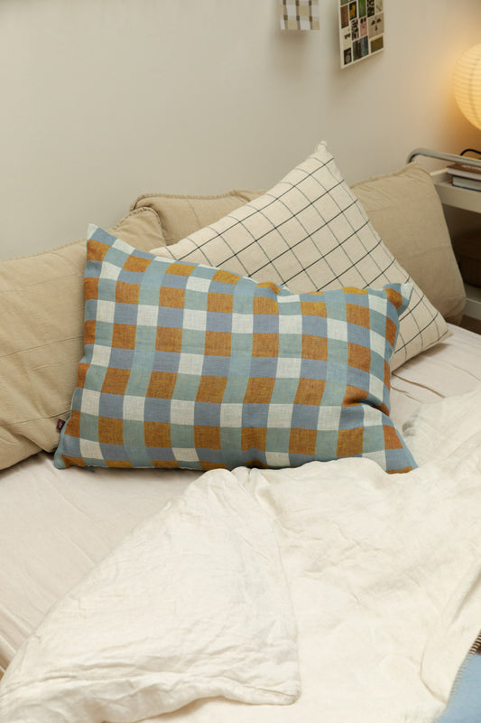Fine Little Day Rutan linen cushion cover in blue and mustard check styled on a bed with House Doctor Join grid check cushion and quilted linen headboard, paper lantern lamp in background.
